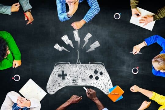 A group of people sit around a chalkboard with a drawing of a game controller.